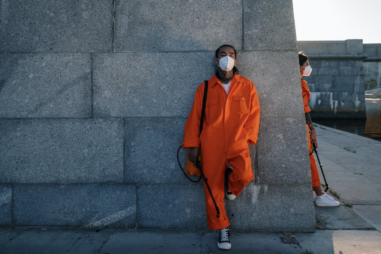 Services Two workers in orange coveralls and facemasks leaning against a wall outdoors.